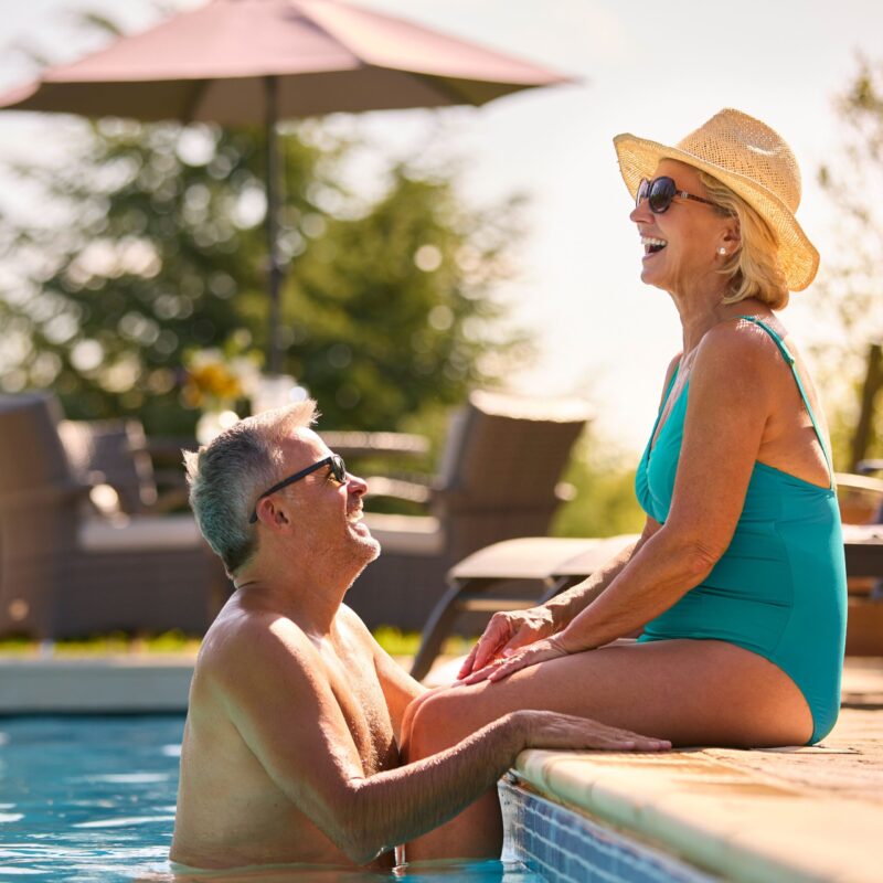 A senior couple enjoy a warm day in the pool. A senior man stands in the pool, smiling up at a senior woman who is sitting on the edge of the pool with her legs in the water. They are both laughing and enjoying the weather.