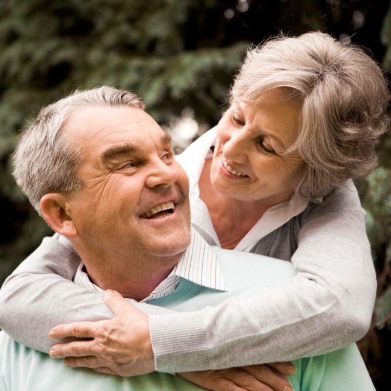 A senior couple embrace outside in the greenery. A senior woman hugs a senior man from behind, smiling down at him.