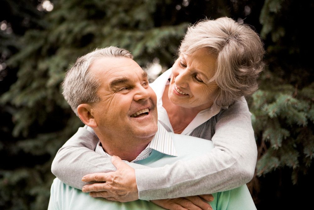 A senior couple embrace outside in the greenery. A senior woman hugs a senior man from behind, smiling down at him.