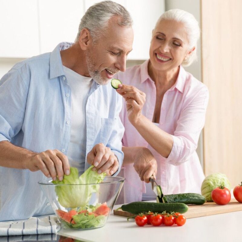 Two seniors smile while making a salad together in the kitchen.