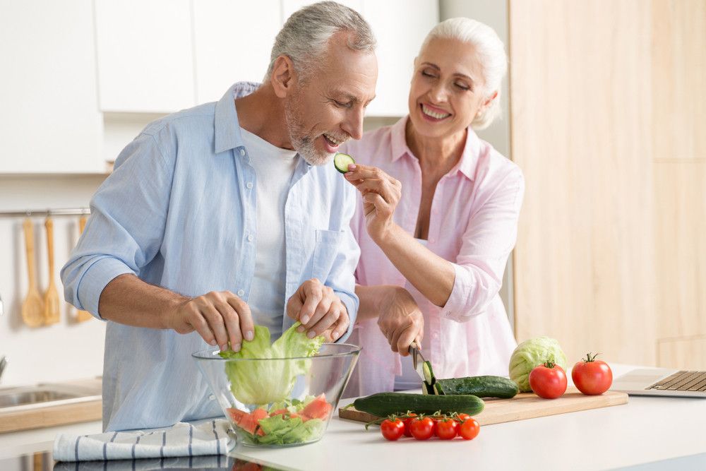 Two seniors smile while making a salad together in the kitchen.
