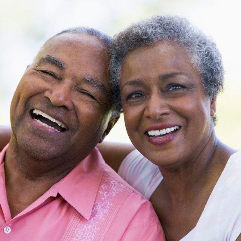 A senior couple sit together, smiling directly at the camera.