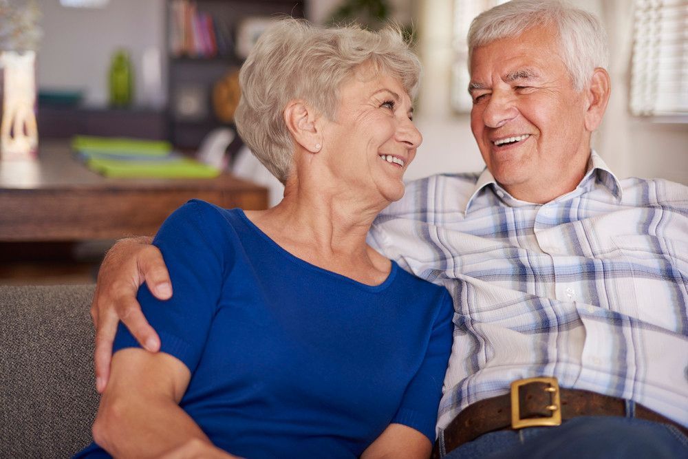 A senior couple sit together on a couch, smiling happily at each other.