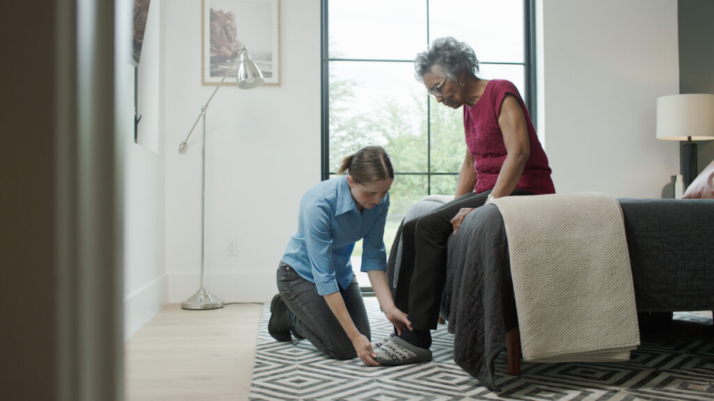 A staff member assists a senior woman with putting on a pair of slippers in her bedroom.