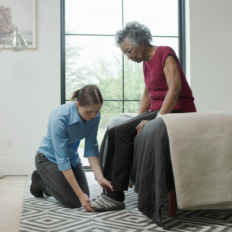 A staff member assists a senior woman with putting on a pair of slippers in her bedroom.