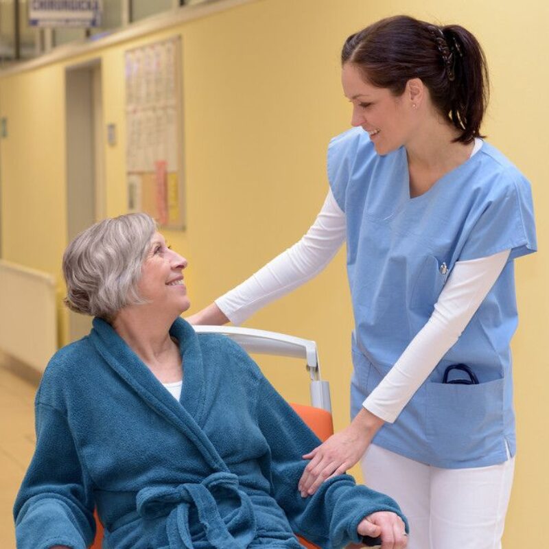 A senior woman receives care from a younger woman in blue and white scrubs. They are smiling at each other.