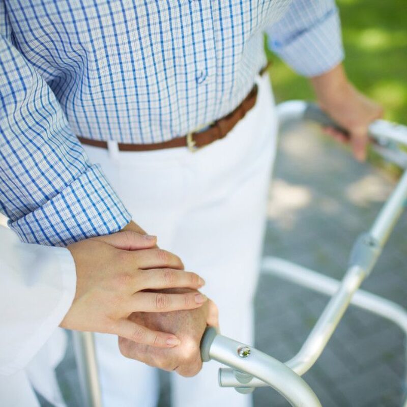 Close up of a staff members hand gently supporting a senior person's hand as they use a walker outside.