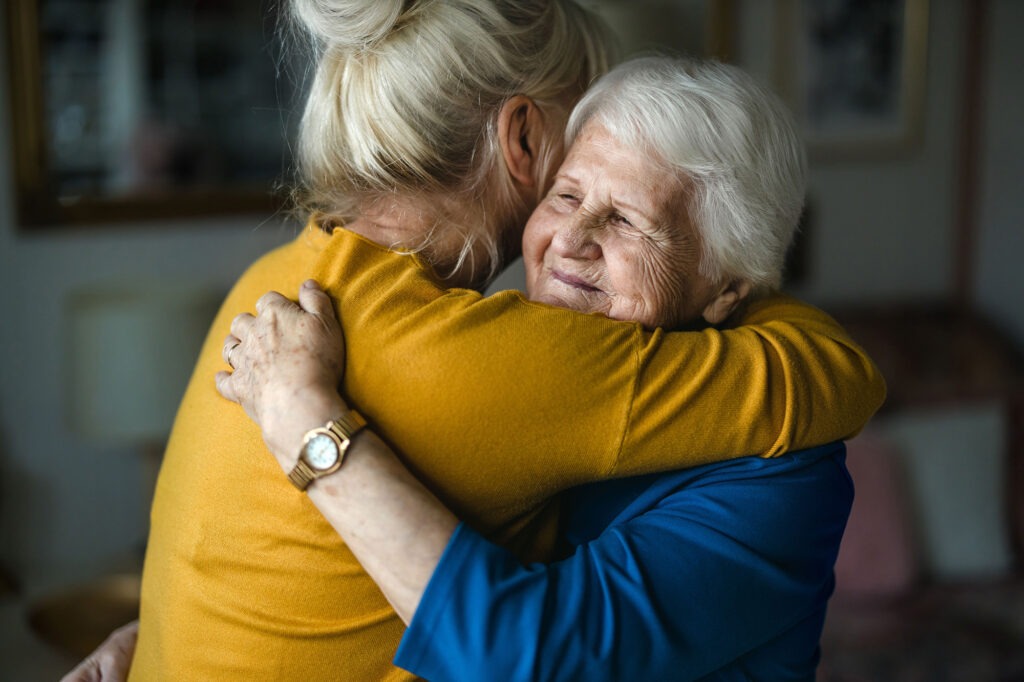 An older woman embraces a younger woman, smiling over her shoulder.