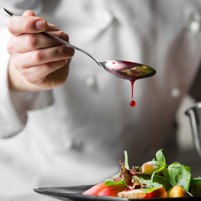 A chef uses a metal spoon to pour red salad dressing over a colorful salad on a black plate.