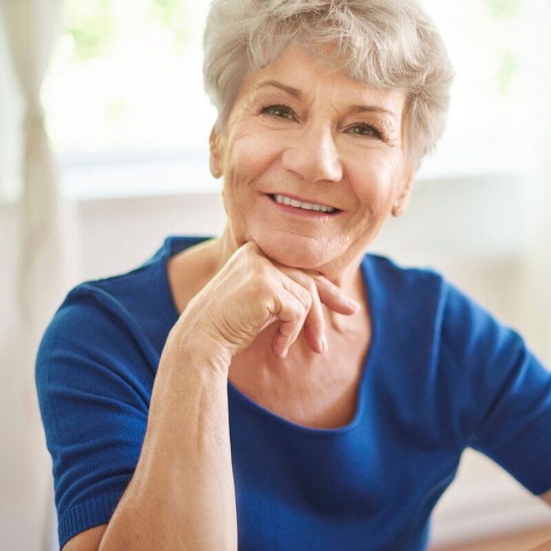 A senior woman rests her chin on her hand and smiles directly into the camera.