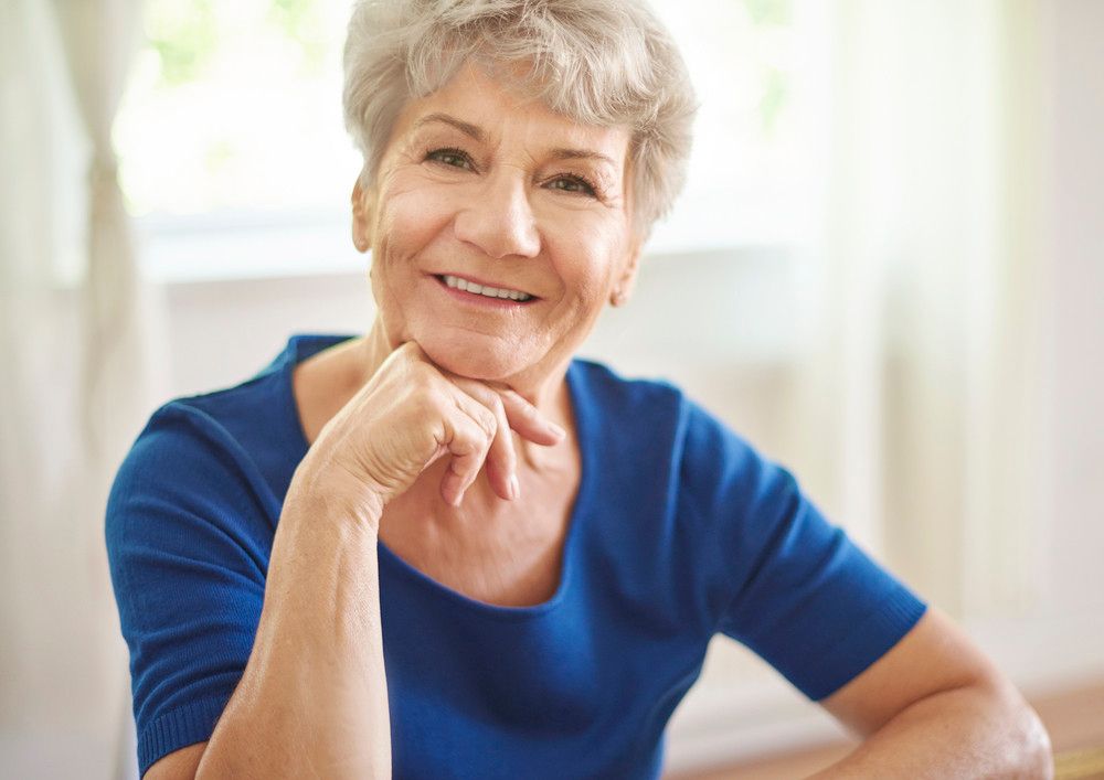 A senior woman rests her chin on her hand and smiles directly into the camera.